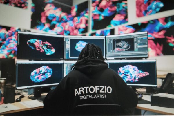 A person wearing a hoodie with "Artofzio" text, looking at multiple screens.