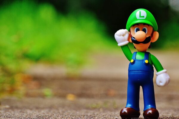 A small Luigi toy figure standing on a wooden surface against a blurry green background.